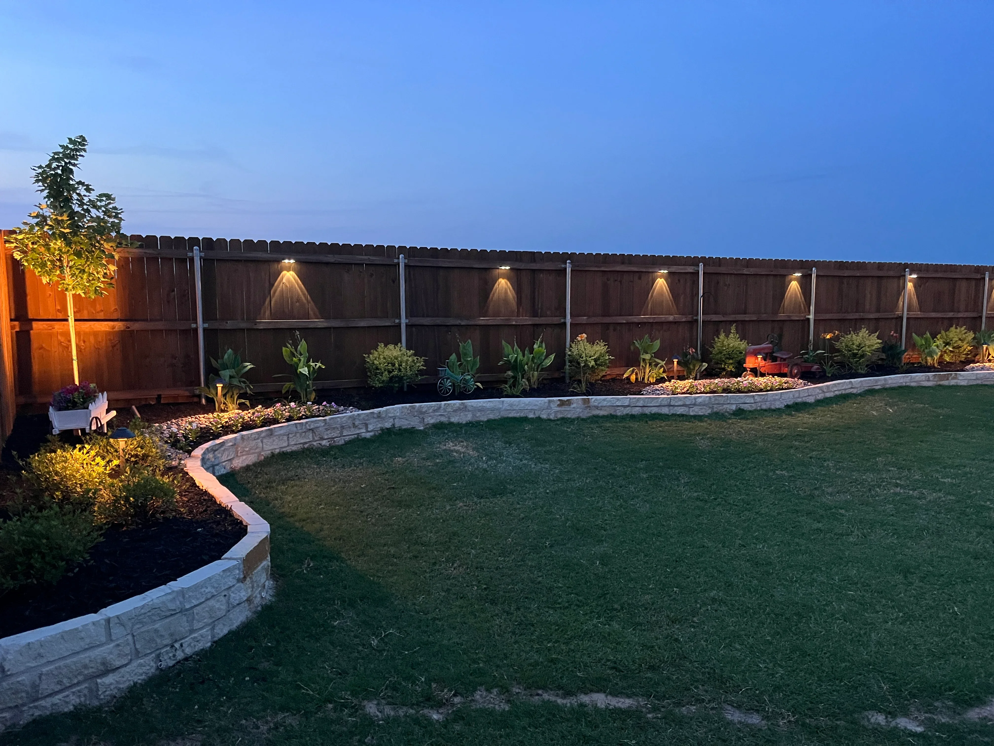 Curved concrete edging, planted beds with ornamentals, landscape lighting, and lawn in backyard setting