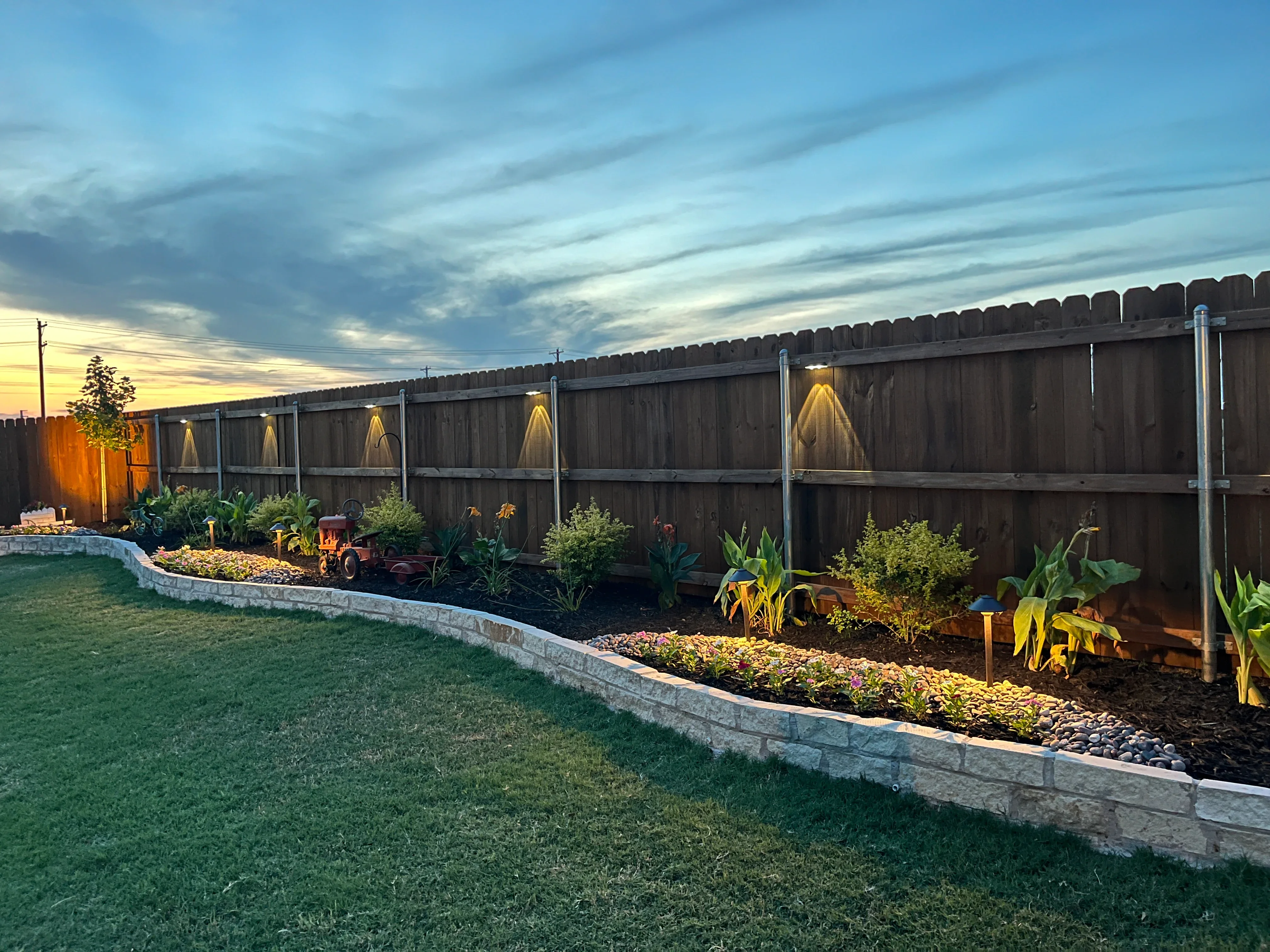 Curved concrete planter beds with mulch, plants, and landscape lighting along wooden fence