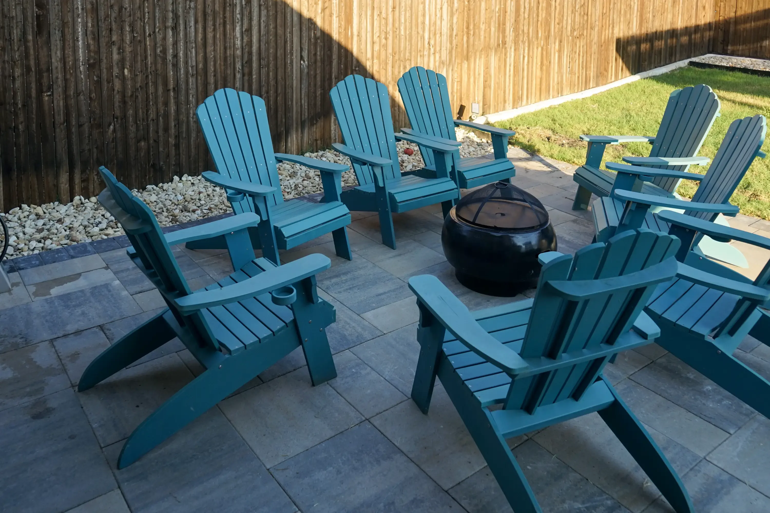 Gray paver patio with blue Adirondack chairs and black fire pit, bordered by wood fence and gravel landscaping