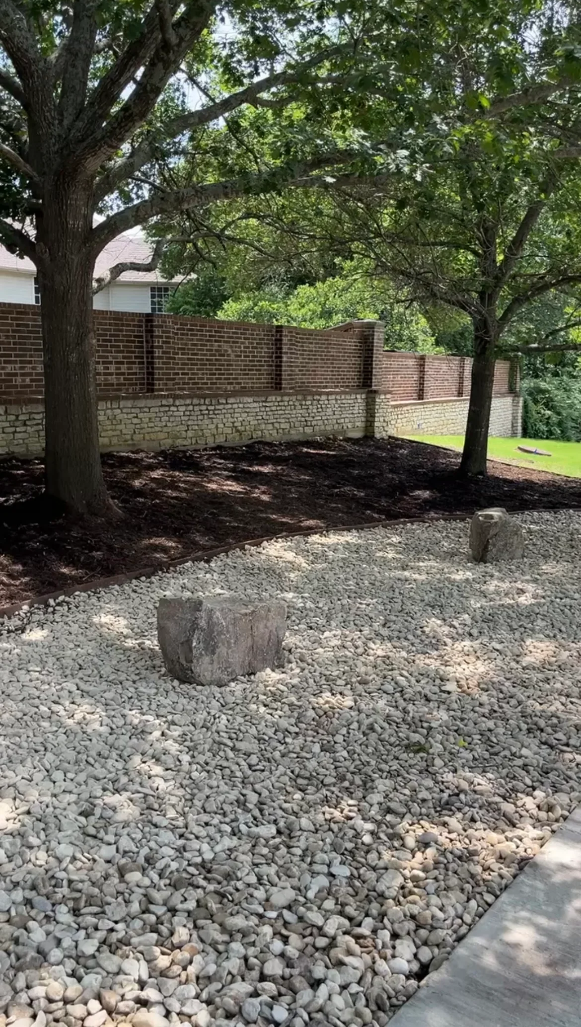 Gravel and mulch landscape with stone edging beneath mature trees against brick wall