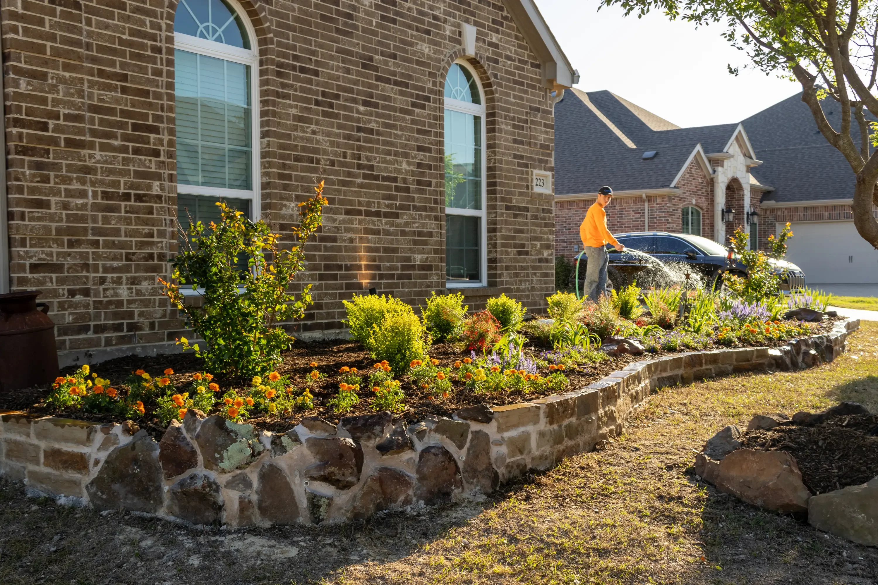 Stone-edged landscape bed with mixed shrubs, yellow flowers, and mulch along brick home foundation