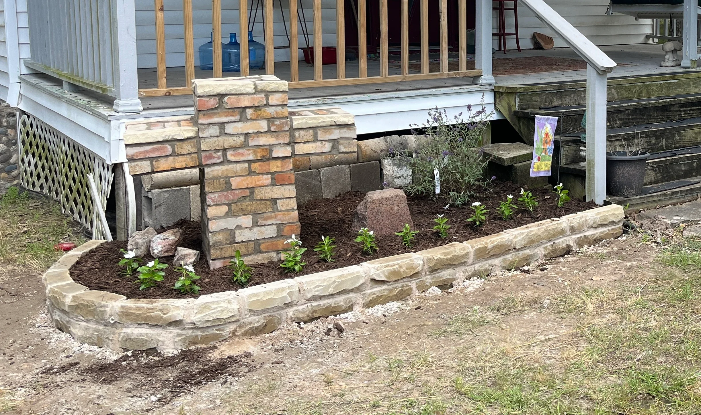 Curved stone-edged planting bed with brick foundation supports and newly planted shrubs in dark mulch